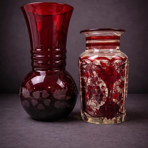 Op de afbeelding: Twee antieke glazen vazen. De vaas links is dieprood met een bolvormige basis en een uitlopende bovenkant, met geëtste bladeren. De vaas rechts is helder met rood geschilderde bloemen- en druivenontwerpen.