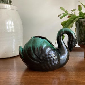 May include: A ceramic swan-shaped planter featuring a glossy finish with a deep black base that transitions into a vibrant teal green glaze on the upper body and neck. The swan has a curved neck and a textured wing design on its side.