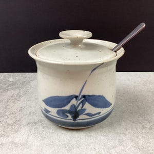 Peut inclure: Un pot en céramique avec un couvercle et une cuillère. Le pot est blanc cassé avec des motifs floraux bleus et un couvercle assorti. Une cuillère en acier inoxydable repose sur le bord. Le pot est probablement destiné à la cuisine.