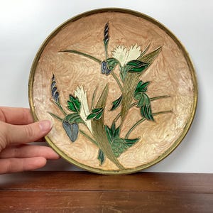 Puede incluir: Un plato decorativo redondo con un dise&ntilde;o floral. El plato tiene un fondo marr&oacute;n claro y un borde dorado. El dise&ntilde;o floral presenta flores blancas, hojas verdes y detalles azules.