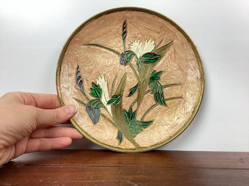 Puede incluir: Un plato decorativo redondo con un dise&ntilde;o floral. El plato tiene un fondo marr&oacute;n claro y un borde dorado. El dise&ntilde;o floral presenta flores blancas, hojas verdes y detalles azules.