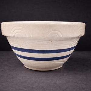 Puede incluir: Un cuenco de cerámica color crema con dos rayas azules horizontales. El cuenco tiene un borde ancho con un patrón decorativo y una superficie texturizada. Este cuenco vintage es adecuado para la cocina.