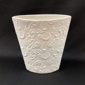 May include: A white ceramic vase with a swirling, embossed pattern. The vase has a trapezoidal shape, wider at the top and narrower at the base. The decorative design covers the entire surface of the vase.