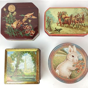 Puede incluir: Cuatro cajas de hojalata vintage con diferentes diseños. Una caja presenta un fondo rojo con dos patos volando sobre un campo verde. Otra caja tiene un fondo rojo con un carruaje tirado por caballos y personas en el fondo. La tercera caja tiene un fondo verde con una iglesia en la distancia. La cuarta caja tiene un fondo azul con un conejo blanco sosteniendo flores.