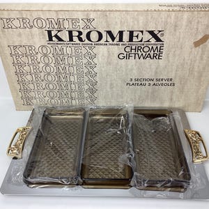 May include: Chrome serving tray with three sections, each with a clear, textured glass insert. The tray is made of silver metal and has gold-colored handles with a floral design. The box it came in is visible in the background, with the brand name "KROMEX" and the text "Chrome Giftware" printed on it.