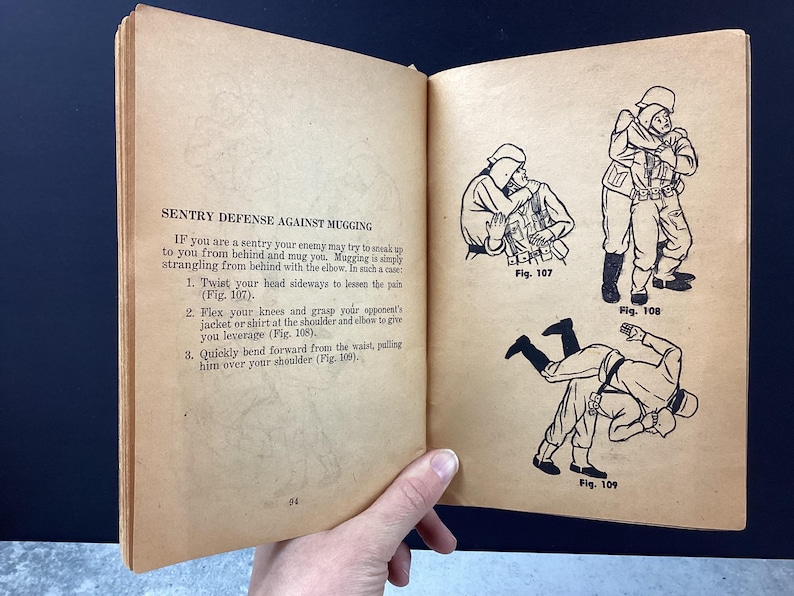 May include: Open book page with text and illustrations about sentry defense against mugging. The text describes techniques, accompanied by three black and white illustrations showing the steps. The book is held open by a hand.