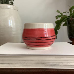 Peut inclure: Un petit bol en céramique à la finition brillante, présentant une partie supérieure de couleur crème et une partie inférieure rose corail vif, orné de plusieurs fines lignes noires horizontales peintes à la main sur la surface.