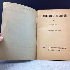 May include: Open book titled "Lightning Ju-Jitsu" by Harry Lord, illustrated by Allen Pickens. The book is published by Technical Publishing Company, Toronto, ONT. The pages are aged with a light brown hue.