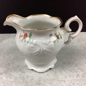May include: White porcelain creamer with a gold rim and a decorative handle. The creamer features embossed floral designs and hand-painted flowers in pink, purple, and green. A classic piece for serving cream or milk.