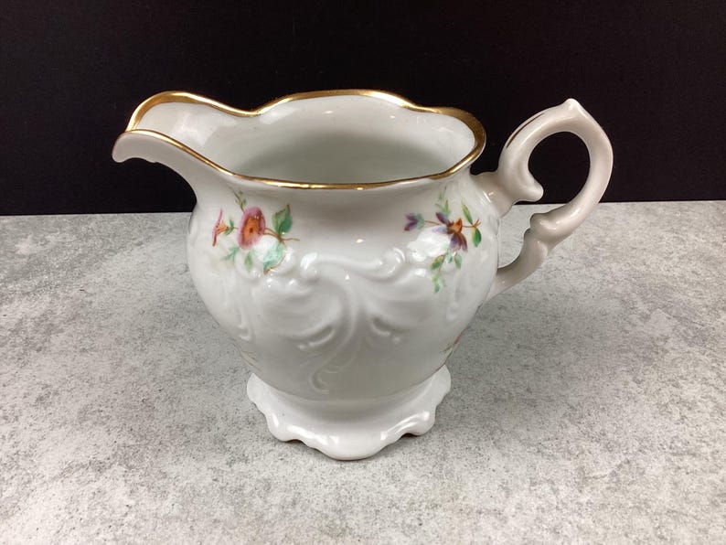 May include: White porcelain creamer with a gold rim and a decorative handle. The creamer features embossed floral designs and hand-painted flowers in pink, purple, and green. A classic piece for serving cream or milk.