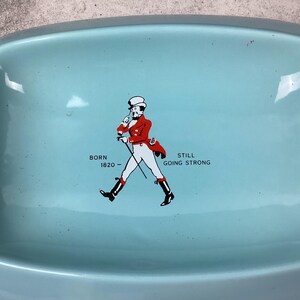 May include: A pale blue rectangular tray with curved edges. It displays a graphic of a man in a red coat and black boots, accompanied by the text "BORN 1820 - STILL GOING STRONG".