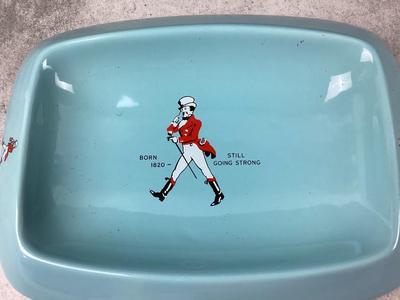 May include: A pale blue rectangular tray with curved edges. It displays a graphic of a man in a red coat and black boots, accompanied by the text "BORN 1820 - STILL GOING STRONG".