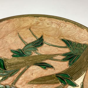 Puede incluir: Primer plano de un objeto decorativo redondo con un borde dorado. El objeto presenta un dise&ntilde;o floral con hojas y tallos verdes sobre un fondo color melocot&oacute;n. El dise&ntilde;o es detallado e intrincado.