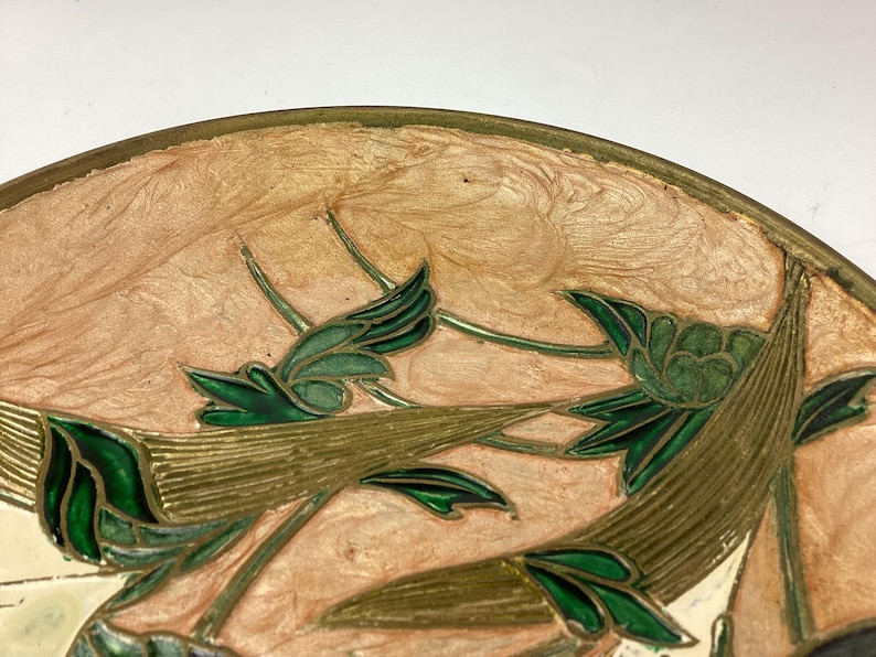 Puede incluir: Primer plano de un objeto decorativo redondo con un borde dorado. El objeto presenta un dise&ntilde;o floral con hojas y tallos verdes sobre un fondo color melocot&oacute;n. El dise&ntilde;o es detallado e intrincado.