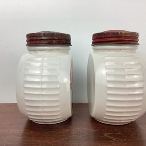 以下が含まれることがあります： 赤と銀色の金属製の蓋が付いた、2つのヴィンテージの白いガラス製スパイスジャー。ジャーの側面にはリブのデザインがあります。ジャーは木製の表面にあります。