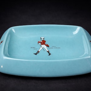 May include: A pale blue ceramic ashtray with a rectangular shape and curved edges. It features a graphic of a man in a red coat and the text "BORN 1820 - STILL GOING STRONG". The ashtray has a glossy finish and is set against a dark background.