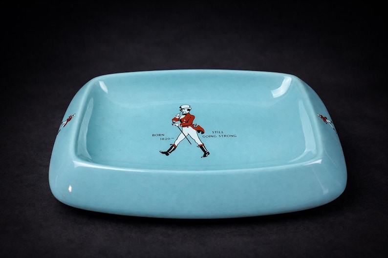 May include: A pale blue ceramic ashtray with a rectangular shape and curved edges. It features a graphic of a man in a red coat and the text "BORN 1820 - STILL GOING STRONG". The ashtray has a glossy finish and is set against a dark background.