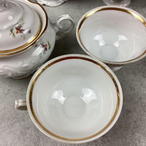 May include: White porcelain teacups and a teapot with gold trim and floral accents. The teacups have handles and a gold rim. The teapot has a lid and a handle. The set is on a gray surface.