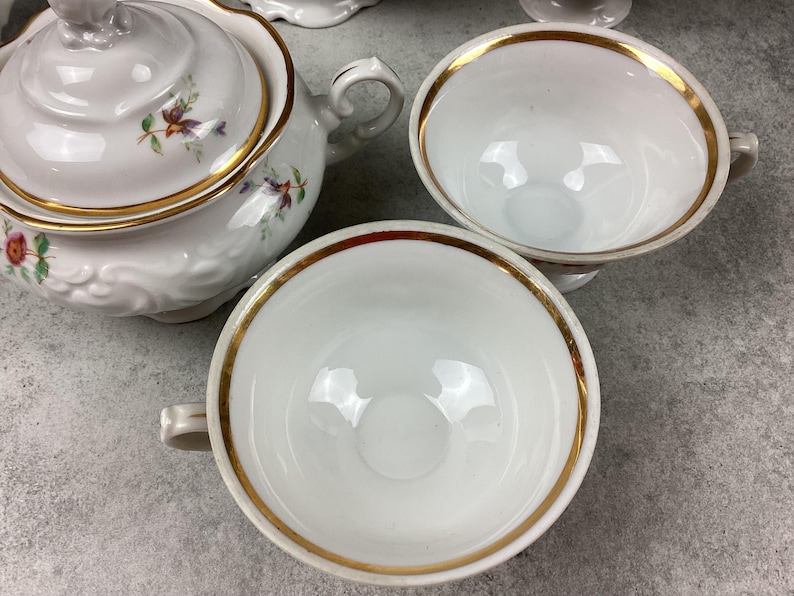May include: White porcelain teacups and a teapot with gold trim and floral accents. The teacups have handles and a gold rim. The teapot has a lid and a handle. The set is on a gray surface.
