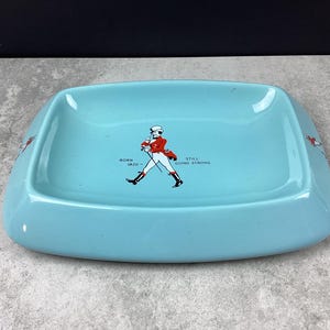 May include: A turquoise rectangular ashtray with rounded corners. The ashtray features a graphic of a man in a red coat and the text "BORN 1820 STILL GOING STRONG". The ashtray has a glossy finish.