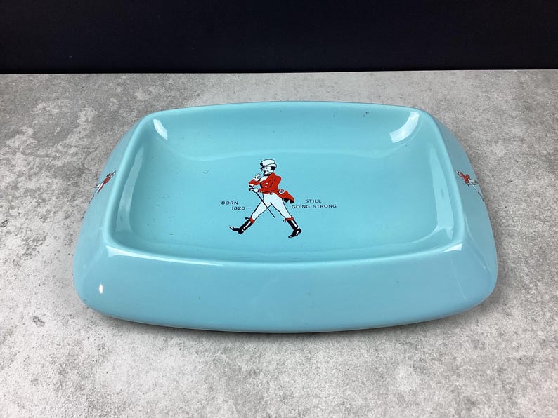 May include: A turquoise rectangular ashtray with rounded corners. The ashtray features a graphic of a man in a red coat and the text "BORN 1820 STILL GOING STRONG". The ashtray has a glossy finish.
