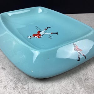May include: A vintage, light blue ashtray with a unique, rounded rectangular shape. The ashtray features two illustrations of skiers in red and white attire. One illustration shows a skier mid-fall, and the other shows a skier walking.