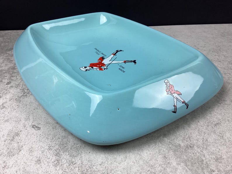 May include: A vintage, light blue ashtray with a unique, rounded rectangular shape. The ashtray features two illustrations of skiers in red and white attire. One illustration shows a skier mid-fall, and the other shows a skier walking.
