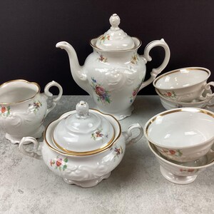 May include: A white porcelain tea set with floral accents. The set includes a teapot, sugar bowl, creamer, and two teacups with saucers. Gold trim edges the cups and sugar bowl. The teapot has a decorative lid.