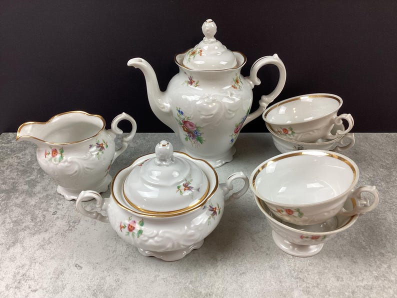May include: A white porcelain tea set with floral accents. The set includes a teapot, sugar bowl, creamer, and two teacups with saucers. Gold trim edges the cups and sugar bowl. The teapot has a decorative lid.