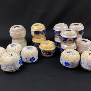May include: A collection of fifteen round balls of crochet thread in various shades of cream, beige, and light yellow. Each ball is wrapped with a blue and white paper label featuring the brand names Coats Opera or DMC Cebelia.