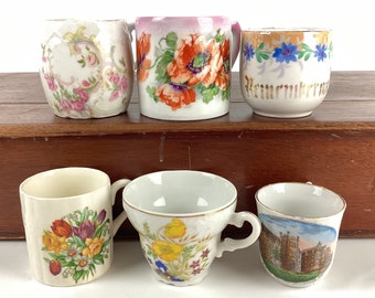 Può includere: Una collezione di sei tazze e tazze antiche. Le tazze presentano disegni floreali in vari colori, tra cui rosa, rosso, giallo e blu. Alcune tazze hanno finiture dorate e motivi decorativi. Una tazza ha un'immagine panoramica.