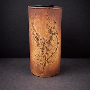 May include: A cylindrical ceramic vase with a textured, brown finish. The vase has an embossed botanical design of a flowering plant. The interior is dark, and the vase stands on a dark surface against a black background.