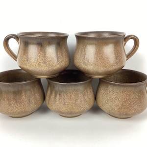 May include: Five brown ceramic mugs with handles are stacked. The mugs have a speckled texture, with a darker brown rim and handle. These mugs are likely for tea or coffee and are stacked in a pyramid.