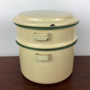 Puede incluir: Contenedor de alimentos de esmalte de dos niveles de color crema con tapa. El contenedor tiene un borde verde y dos asas en cada sección. La tapa tiene un pequeño desconchado. Un artículo de cocina vintage.