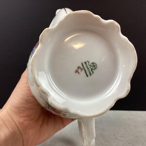 May include: White porcelain creamer with a scalloped rim and a handle. The bottom of the creamer features a green and red stamp with the text "Bavaria" and the number "72". The creamer has floral designs in various colors.