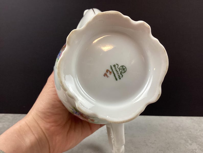 May include: White porcelain creamer with a scalloped rim and a handle. The bottom of the creamer features a green and red stamp with the text "Bavaria" and the number "72". The creamer has floral designs in various colors.