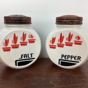 Puede incluir: Un par de saleros y pimenteros blancos vintage. Cada salero tiene un diseño rojo y negro de plantas en macetas. Las tapas son de metal con un acabado rojo. Las palabras "SALT" y "PEPPER" están impresas.