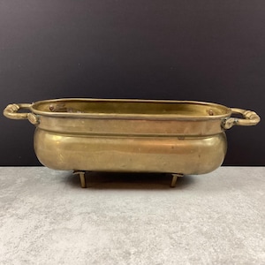 Peut inclure: Un pot de fleurs en laiton vintage, de forme rectangulaire avec des côtés incurvés et deux poignées ornées. Il repose sur quatre petits pieds. Le pot a une teinte dorée chaude et montre des signes d'âge.
