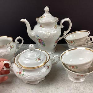 May include: A white porcelain tea set with floral accents and gold trim. The set includes a teapot, sugar bowl, creamer, and two teacups with saucers. The teapot has a decorative lid and a curved spout. The teacups are stacked.