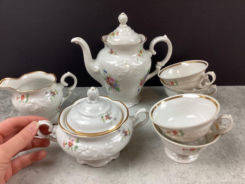 May include: A white porcelain tea set with floral accents and gold trim. The set includes a teapot, sugar bowl, creamer, and two teacups with saucers. The teapot has a decorative lid and a curved spout. The teacups are stacked.
