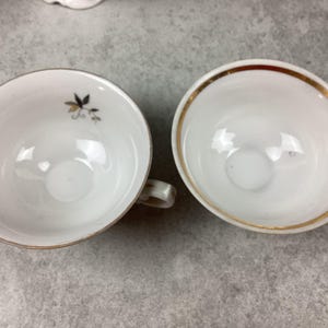 May include: Two white teacups with gold trim and small handles. One cup has a small floral design in black and gold. The cups are empty and sit on a gray surface. The cups are likely part of a tea set.