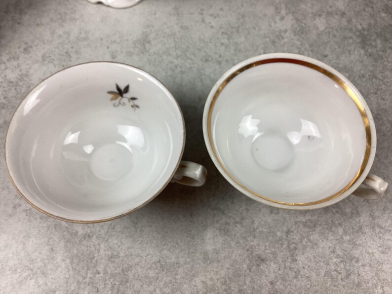 May include: Two white teacups with gold trim and small handles. One cup has a small floral design in black and gold. The cups are empty and sit on a gray surface. The cups are likely part of a tea set.