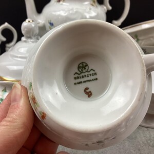 May include: White porcelain teacup with a floral design and gold trim. The teacup is part of a tea set, with other pieces visible in the background. The bottom of the teacup has the text "WAŁBRZYCH MADE IN POLAND".
