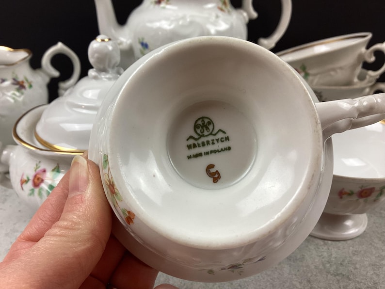 May include: White porcelain teacup with a floral design and gold trim. The teacup is part of a tea set, with other pieces visible in the background. The bottom of the teacup has the text "WAŁBRZYCH MADE IN POLAND".