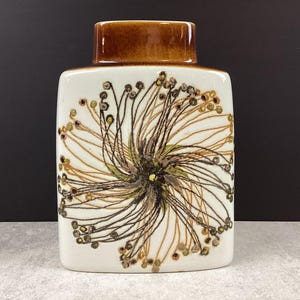 May include: A rectangular ceramic vase with a brown top and a white body. The body features a detailed floral design in brown, tan, and green. The vase has a square shape and a smooth finish, suitable for home decor.