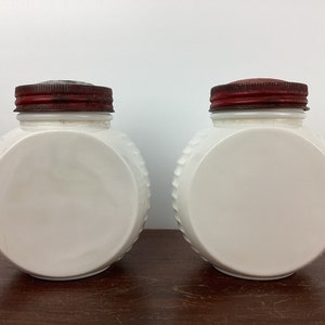 以下が含まれることがあります： テクスチャ加工された側面と前面に大きな滑らかな円がある、2つの白い丸い瓶。各瓶には、錆びた赤色の金属製の蓋が付いています。瓶は濃い木製の表面に表示されています。