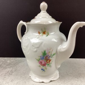 May include: White porcelain teapot with a lid and a curved spout. The teapot features a floral design with pink, blue, and orange flowers. The handle and spout are curved. The teapot is on a white base and has a gold trim.