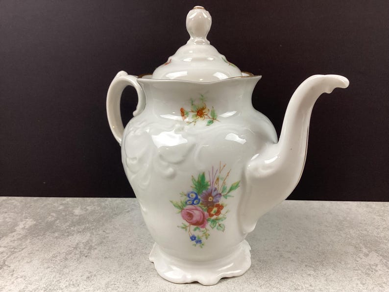 May include: White porcelain teapot with a lid and a curved spout. The teapot features a floral design with pink, blue, and orange flowers. The handle and spout are curved. The teapot is on a white base and has a gold trim.