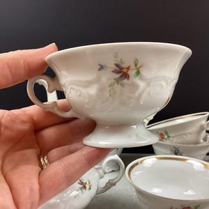 May include: White porcelain teacups with floral designs and gold trim. The teacups have ornate handles and are set on a gray surface. One teacup is held by a hand, and several others are stacked in the background.