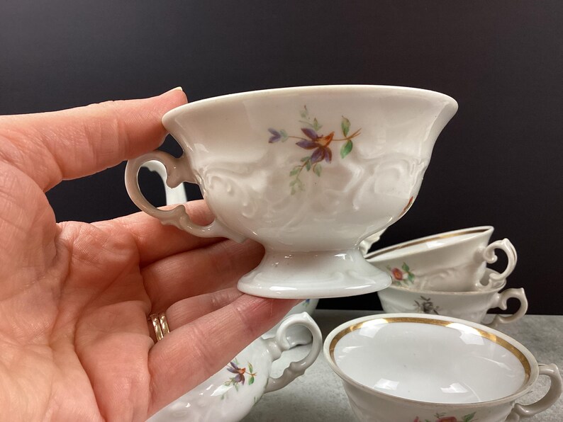 May include: White porcelain teacups with floral designs and gold trim. The teacups have ornate handles and are set on a gray surface. One teacup is held by a hand, and several others are stacked in the background.
